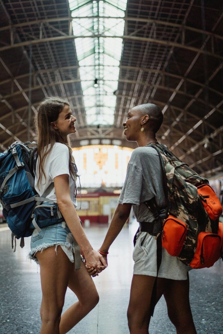 Two women holding hands with backpacks at a train station, expressing love.